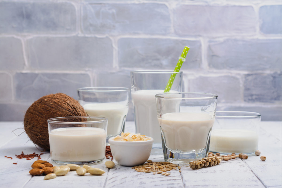 Eine Auswahl verschiedener Pflanzenmilch in Gläsern, dazu eine Kokosnuss und Nüsse. Wesentliche vegane Backzutaten.  