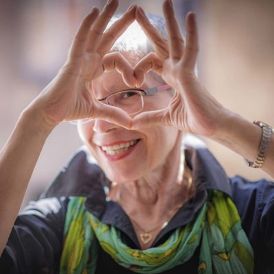 An older woman making a love heart shape with her fingers and smiling. 