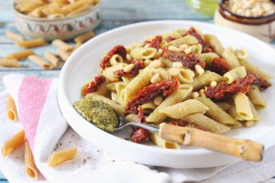 a white bowl is filled with penne pasta, sundried tomatoes, and pine nuts with a bamboo handled spoon of creamy pesto