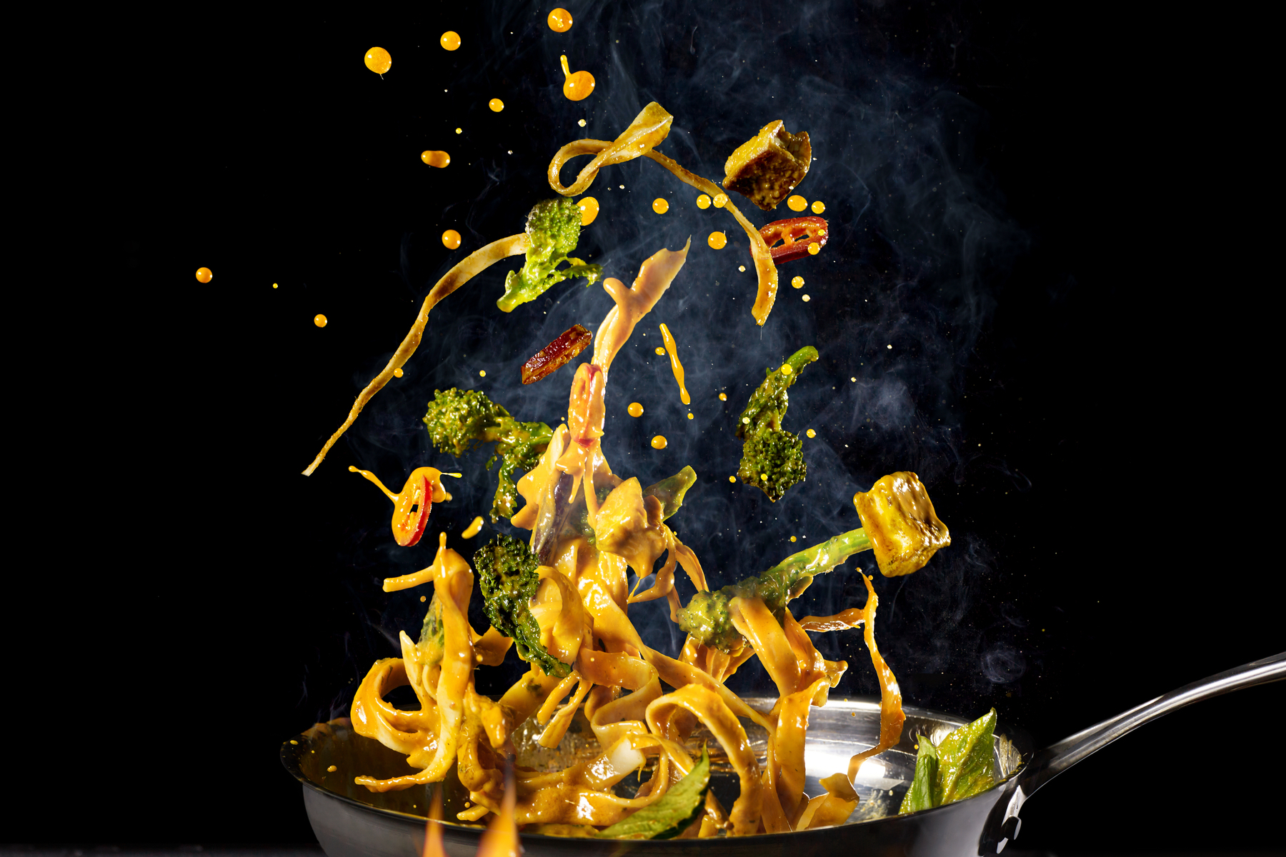 Fresh veggies, noodles, and sauce are tossed in the air by a black skillet