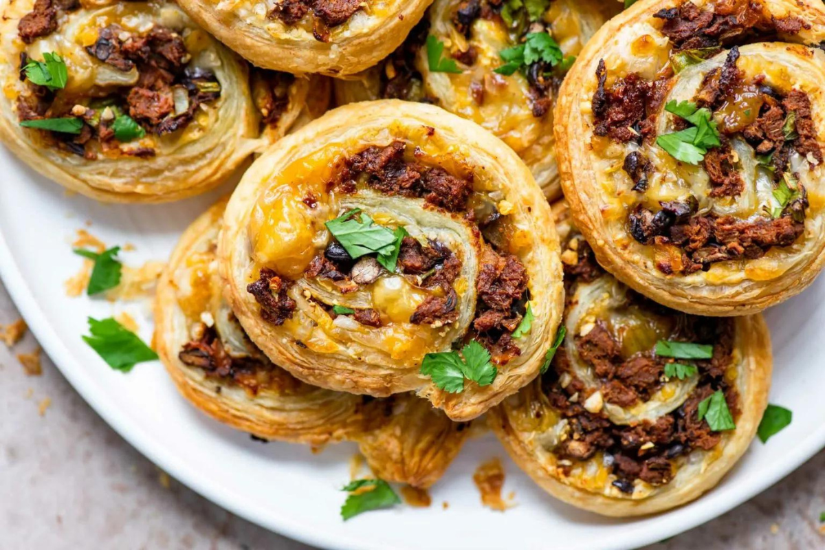 Delicious golden doughy pinwheels stuffed with vegan meat and cheese sit piled on a white plate and are garnished with cilantro