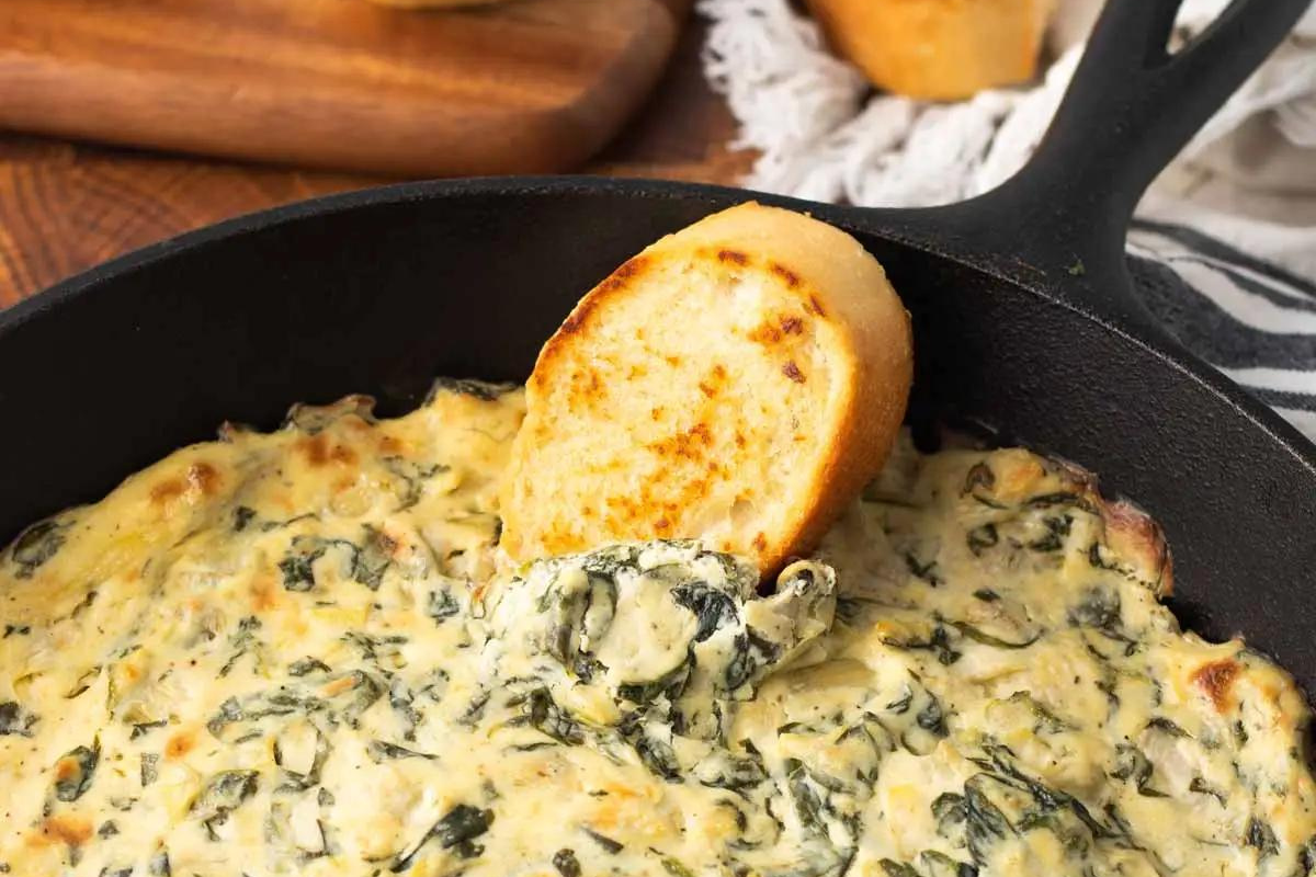 A piece of golden crispy baguette is dipped in a cast iron skillet of spinach and artichoke dip 