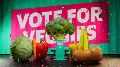 Vegetables "stand" at a podium with a banner behind them that states "vote for veggies"